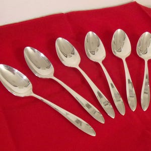 May include: Six silver spoons arranged on a red surface. The spoons have oval bowls and long, slender handles. The spoons are reflective, and the red surface is slightly textured.