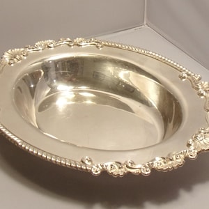 May include: Oval silver serving bowl with ornate floral and rope detailing around the rim. The interior of the bowl is highly reflective, and the piece is set against a neutral background. A decorative piece for serving.