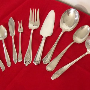 May include: An assortment of antique silver serving utensils, including spoons, forks, and a cake server, arranged on a vibrant red tablecloth. The silverware gleams, reflecting light, and showcases intricate designs.