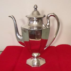 May include: A silver-colored, Art Deco-style coffee pot with a lid and a curved handle. The pot has a geometric design with a hexagonal base and a spout. It sits on a red surface.