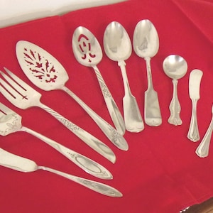 May include: A collection of antique silver serving utensils, including forks, spoons, and butter knives, arranged on a red tablecloth. The utensils have decorative handles and intricate designs, showcasing a vintage aesthetic.
