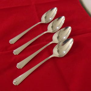 May include: Four silver spoons with elongated handles and oval bowls are arranged diagonally on a red surface. The spoons have a decorative design at the base of the handle. The spoons are reflective.