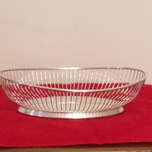 May include: An oval-shaped silver-colored metal basket featuring a wire-frame design with vertical slats. The basket has a solid, smooth base and an open top, constructed from thin, reflective metallic rods that form a continuous, airy structure.