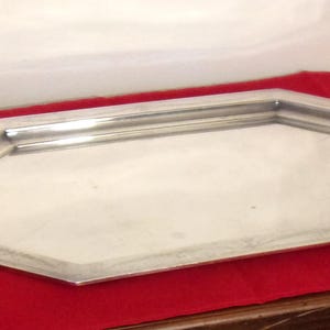 May include: A silver-colored, octagonal serving tray with two handles. The tray is placed on a red surface and has a reflective surface. The handles are curved and add a decorative element to the tray.