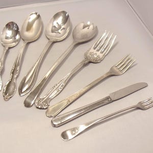 May include: A collection of silver-colored silverware, including spoons, forks, and a knife, arranged on a light-colored surface. The spoons have varying designs, and the forks and knife have simple, elegant handles. The silverware is likely for dining.