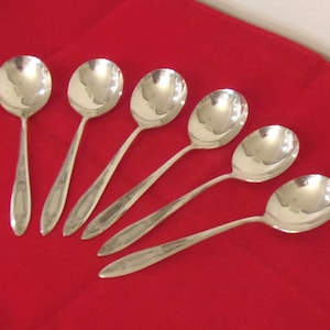 May include: Six silver soup spoons arranged on a vibrant red surface. The spoons have rounded bowls and long, slender handles. The reflective surface of the spoons contrasts with the matte background, creating a visually appealing arrangement.