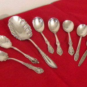 May include: A collection of seven silver-toned metal spoons of varying sizes and handle designs. The set includes one large serving spoon with a scalloped edge, alongside six smaller spoons featuring ornate embossed patterns on their handles.