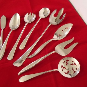 May include: A collection of silver serving utensils, including spoons, forks, and a slotted spoon, arranged on a red surface. The utensils have long handles and are designed for serving food. The set includes a butter knife.