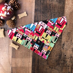 May include: A colorful dog bandana featuring a pattern of nutcracker soldiers in red, blue, green, and gold. The bandana is triangular in shape and has a black background.