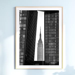 May include: A black and white framed wall art print featuring the Empire State Building centered between two modern glass skyscrapers. The tall, narrow composition highlights the contrast between the historic spire and the dark, grid-patterned facades.