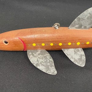 May include: Wooden fish-shaped ornament with a reddish-brown body, silver fins, and yellow dot accents. The fish has a red line near the head and a small metal loop for hanging. A decorative item.