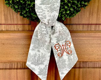 May include: A white fabric bow with a gray toile pattern and an orange embroidered monogram "W". The bow is tied around a green wreath.