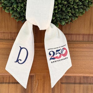 May include: Cream-colored door sash with a navy blue "D" and a 250th Anniversary emblem. The sash is draped over a green wreath, set against a wooden door. The emblem features red, white, and blue colors.