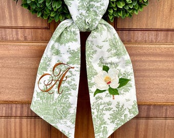 May include: A white and green decorative scarf with a toile pattern and a magnolia flower applique. The scarf has a brown embroidered letter "A" and is draped over a green wreath. The scarf is made of a soft material.