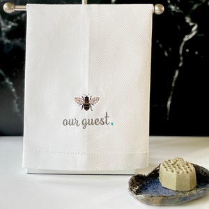 May include: White hand towel with an embroidered bee and the text "our guest."