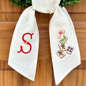 May include: A white linen door swag with a red embroidered letter "S" on one side and a design of mahjong tiles on the other side.