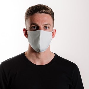 May include: A person wearing a white face mask, looking directly at the camera. The person is wearing a black t-shirt.