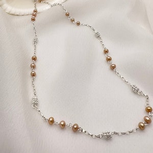 Puede incluir: Un collar de cadena de plata con perlas marrones. La cadena tiene pequeños detalles decorativos de plata.