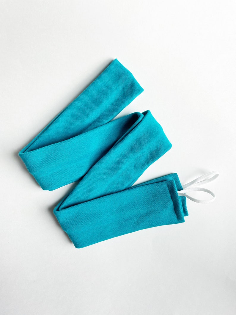 以下が含まれることがあります： 白いゴム紐が付いたターコイズブルーの布製のヘッドバンド。ヘッドバンドは折り畳んで結ばれています。