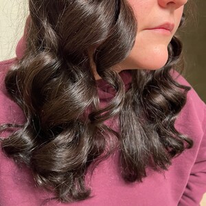 以下が含まれることがあります： 緩やかなカールにスタイリングされた、長く暗い茶色の髪をした女性。彼女は濃い紫色のスウェットシャツを着ています。