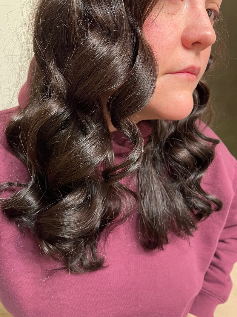 以下が含まれることがあります： 緩やかなカールにスタイリングされた、長く暗い茶色の髪をした女性。彼女は濃い紫色のスウェットシャツを着ています。