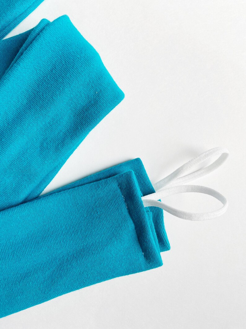 以下が含まれることがあります： 白いゴムひもが付いたターコイズブルーの布製の腕カバー2つ。腕カバーは折り畳まれて、ひもは一緒に結ばれています。