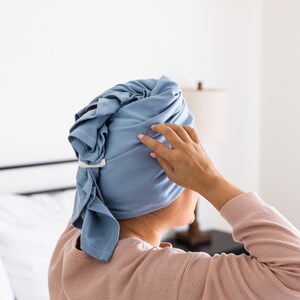 May include: A pale blue hair towel wrap secured with a white clasp. The towel is wrapped around the head. The person is wearing a pink long-sleeved top. The background is a white wall and a bed.