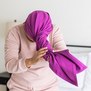 May include: A person wearing a pink waffle-knit long-sleeve shirt and shorts, holding a vibrant purple head covering. The head covering is wrapped and knotted. The background is a white bedroom setting.