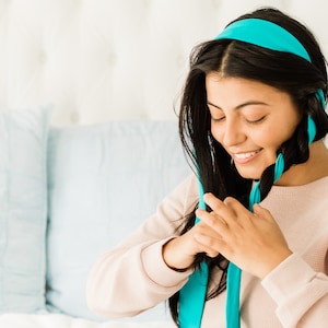 以下が含まれることがあります： 長い黒髪の女性がターコイズブルーのリボンで髪を編んでいます。彼女は薄いピンクのセーターを着ています。