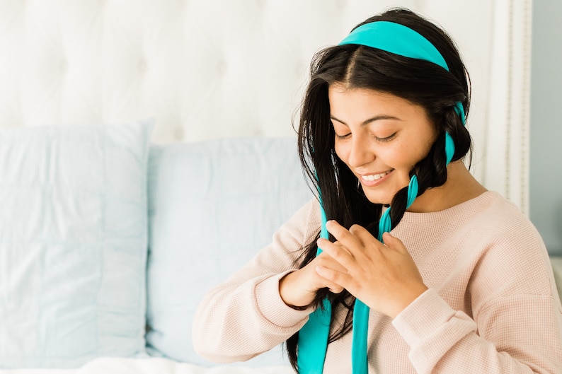 以下が含まれることがあります： 長い黒髪の女性がターコイズブルーのリボンで髪を編んでいます。彼女は薄いピンクのセーターを着ています。