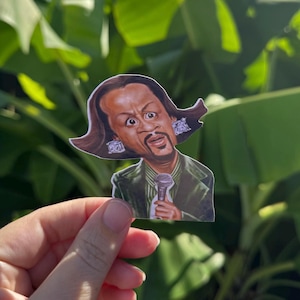 Puede incluir: Una pegatina con una caricatura de un hombre con cabello castaño y perilla, vistiendo un traje verde y sosteniendo un micrófono. El hombre tiene una expresión de sorpresa y lleva pendientes grandes.