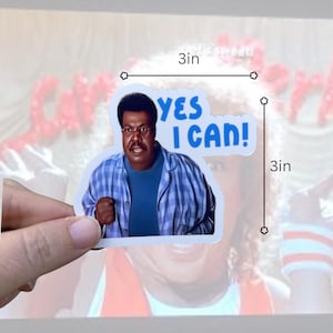 May include: A 3-inch sticker featuring a man with glasses and a mustache, wearing a blue plaid shirt and a blue shirt underneath. The sticker has the text "YES I CAN!" in blue. The sticker is held by a person's hand.