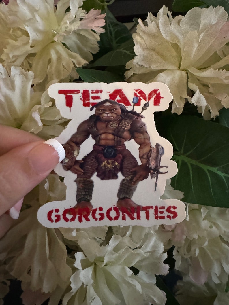 May include: A sticker featuring a muscular, humanoid creature with brown skin and tribal attire. The sticker has the words "TEAM GORGONITES" in red, distressed lettering. The background is a cluster of white flowers.
