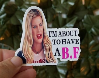 May include: A white sticker featuring a woman with blonde hair and the text "I'M ABOUT TO HAVE A B.F" in black and pink. The sticker is held by a hand against a backdrop of green leaves and fairy lights.