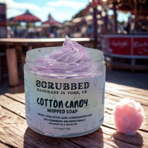 Op de afbeelding: Een pot lavendelkleurige opgeklopte zeep, met het label "Cotton Candy", staat op een houten oppervlak. De pot is gevuld met luchtige zeep en wordt vergezeld door een kleine roze suikerspin. Netto gewicht 100g.