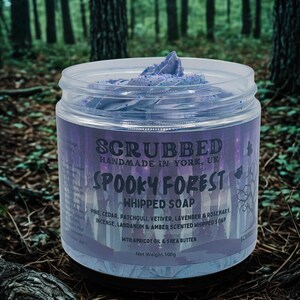 Puede incluir: Un tarro de jabón batido "Spooky Forest" con una etiqueta morada y un fondo de bosque. La etiqueta dice "Scrubbed Handmade in York, UK" y enumera los ingredientes. El jabón batido es de color morado claro y el peso neto es de 100g.