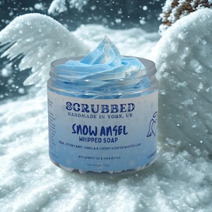 Może przedstawiać: Słoik mydła w piance "Snow Angel" z jasnoniebieską, ubijaną polewą. Słoik ma etykietę z nazwą marki "Scrubbed" i szczegółami zapachu. Tło to zaśnieżona zimowa sceneria.