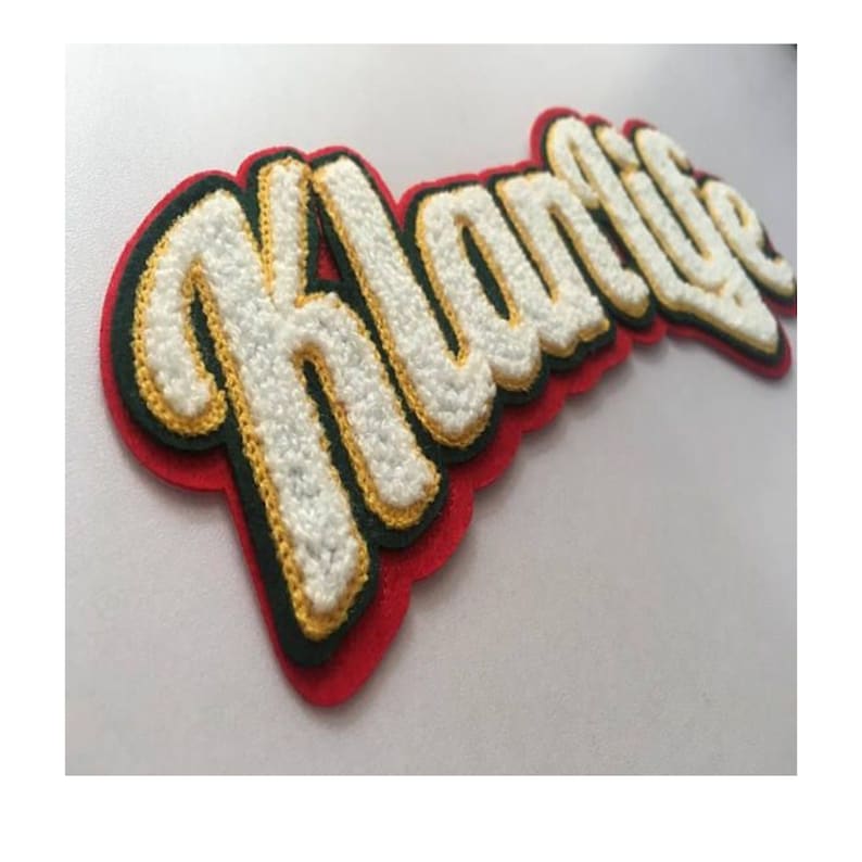 May include: A custom-made patch with the word "Hustle" in a cursive, textured font. The letters are white with a yellow outline, set against a black and red felt background. The patch is designed for apparel.