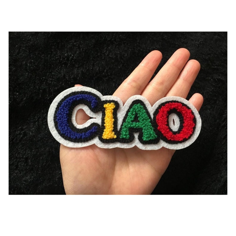 May include: A colorful embroidered patch with the Italian word "CIAO" in a playful, varsity-style font. The letters are in blue, yellow, green, and red, outlined in black and white. The patch is likely for clothing or accessories.