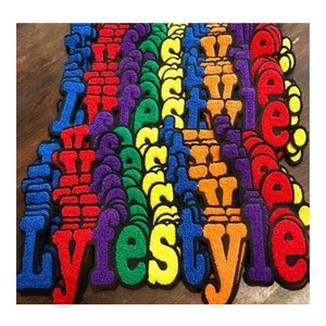 May include: A collection of colorful, embroidered patches spelling out "Lifestyle". The patches feature a variety of vibrant colors, including red, blue, yellow, orange, and purple, with a black border. The letters have a textured, fuzzy appearance.
