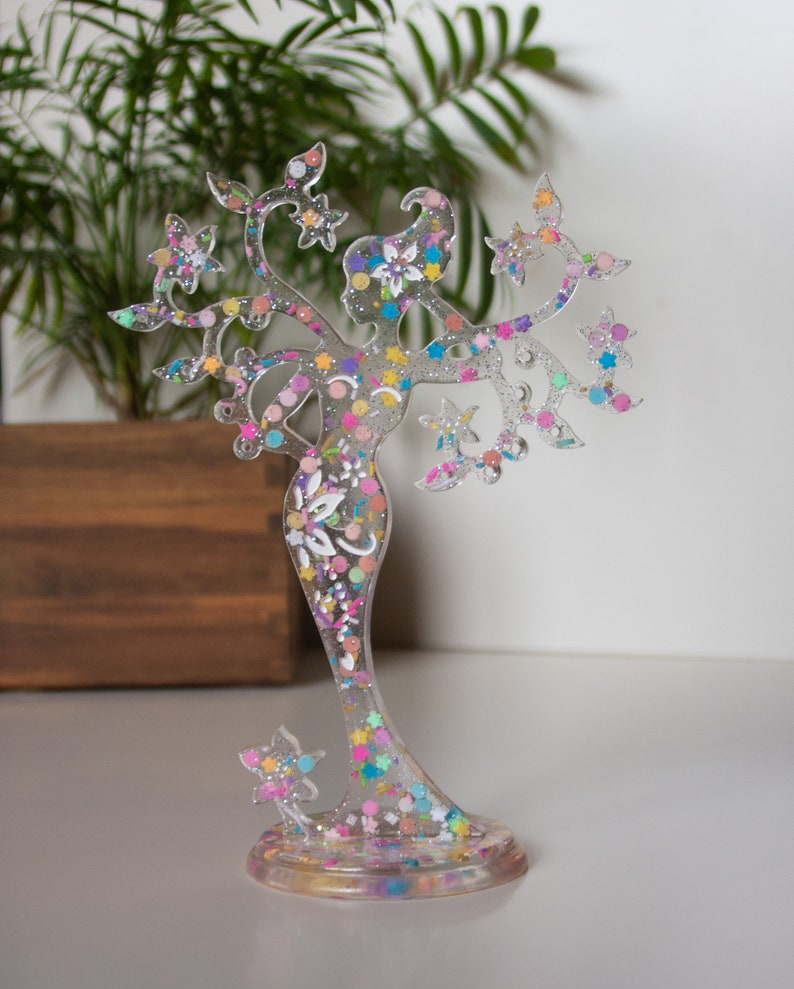 May include: A clear resin figurine of a woman with a tree-like design. The figurine is decorated with colourful glitter and has a base.