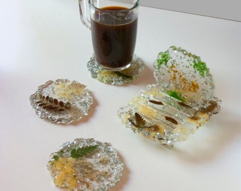 Puede incluir: Cuatro posavasos de vidrio transparente con flores y hojas prensadas en su interior. Los posavasos son redondos y tienen una superficie ligeramente texturizada. Una taza de vidrio de café se encuentra sobre uno de los posavasos.