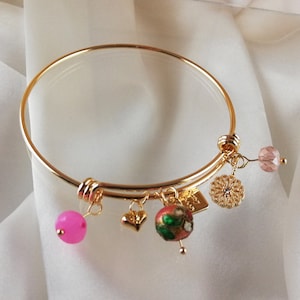 Peut inclure: Un bracelet jonc en or avec plusieurs breloques, dont une perle rose, un charme en forme de cœur, une perle verte et rouge, un charme en or avec un motif floral et une perle de cristal rose.