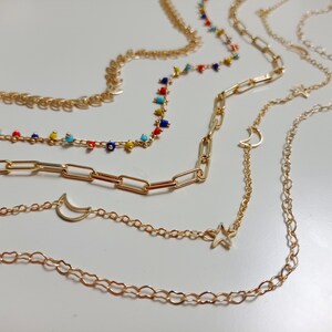 Puede incluir: Cuatro cadenas de oro con diferentes estilos y encantos. Una cadena tiene cuentas de colores, otra tiene un encanto de media luna, otra tiene un encanto de estrella y la última tiene un diseño simple de eslabón de cadena.