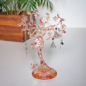 May include: A clear resin jewellery stand shaped like a woman with gold leaf accents and pink and white flowers embedded in the resin. The stand has a round base and is holding necklaces and earrings.
