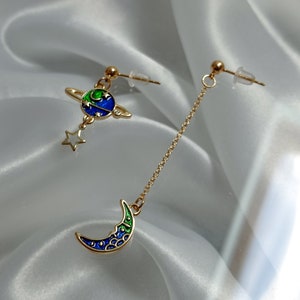 May include: A pair of gold earrings with a celestial theme. One earring features a blue and green planet with a gold ring around it and a small gold star hanging from it. The other earring features a blue and green crescent moon hanging from a gold chain.