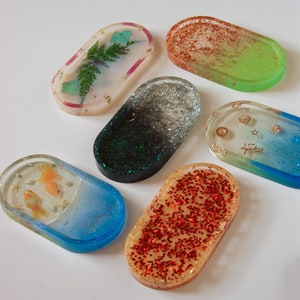 Könnte beinhalten: Sechs ovale Harzschalen mit verschiedenen Designs. Eine Schale hat einen grünen Farn und einen hellblauen Stein, eine andere hat goldenen Glitzer, eine andere hat schwarzen und grünen Glitzer, eine andere hat roten Glitzer, eine andere hat goldenen und blauen Glitzer mit einem Herz- und Sternmuster und eine andere hat einen blauen Hintergrund mit zwei goldenen Fischen.
