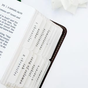May include: Open Bible with white tab dividers labeled with book names. The Bible has a brown leather cover and white pages. The tabs are in a clear plastic material. A white flower and green leaves are in the background.