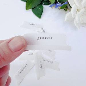May include: A hand holding several white Bible tabs with black text. The tab in focus reads "genesis". Other tabs visible include "1 chronicles", "2 kings", "hose", "nehemiah", "esther", and "haggai".