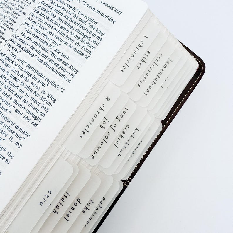 May include: Open Bible with white tab dividers labeled with book names. The pages are open to the Old Testament, with text visible. The Bible has a brown leather cover with white stitching.